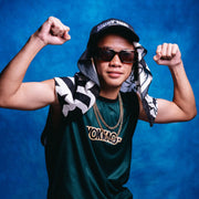 Person flexing muscles wearing a green 'Vonnie' shirt, black cap, and sunglasses against a blue background