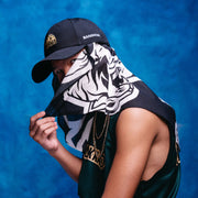 Person wearing a black cap and green sleeveless shirt with a bandana over their face against a blue background