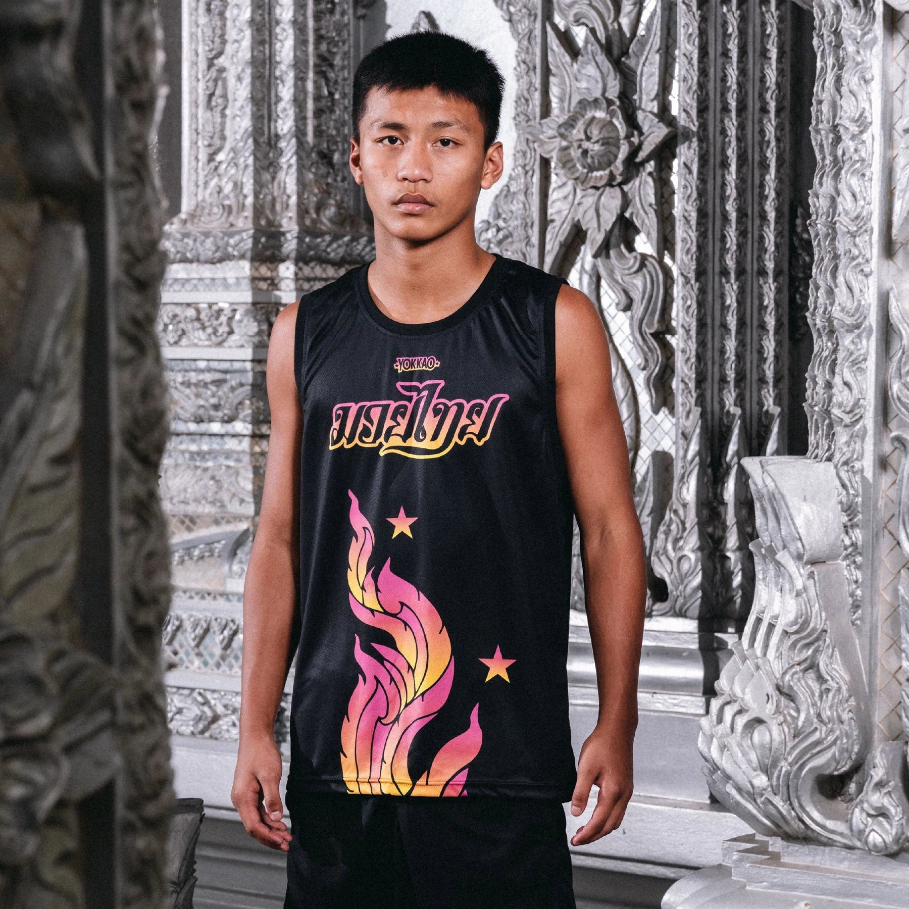 Person wearing a black sleeveless shirt with colorful designs and text, standing in an ornate indoor setting.