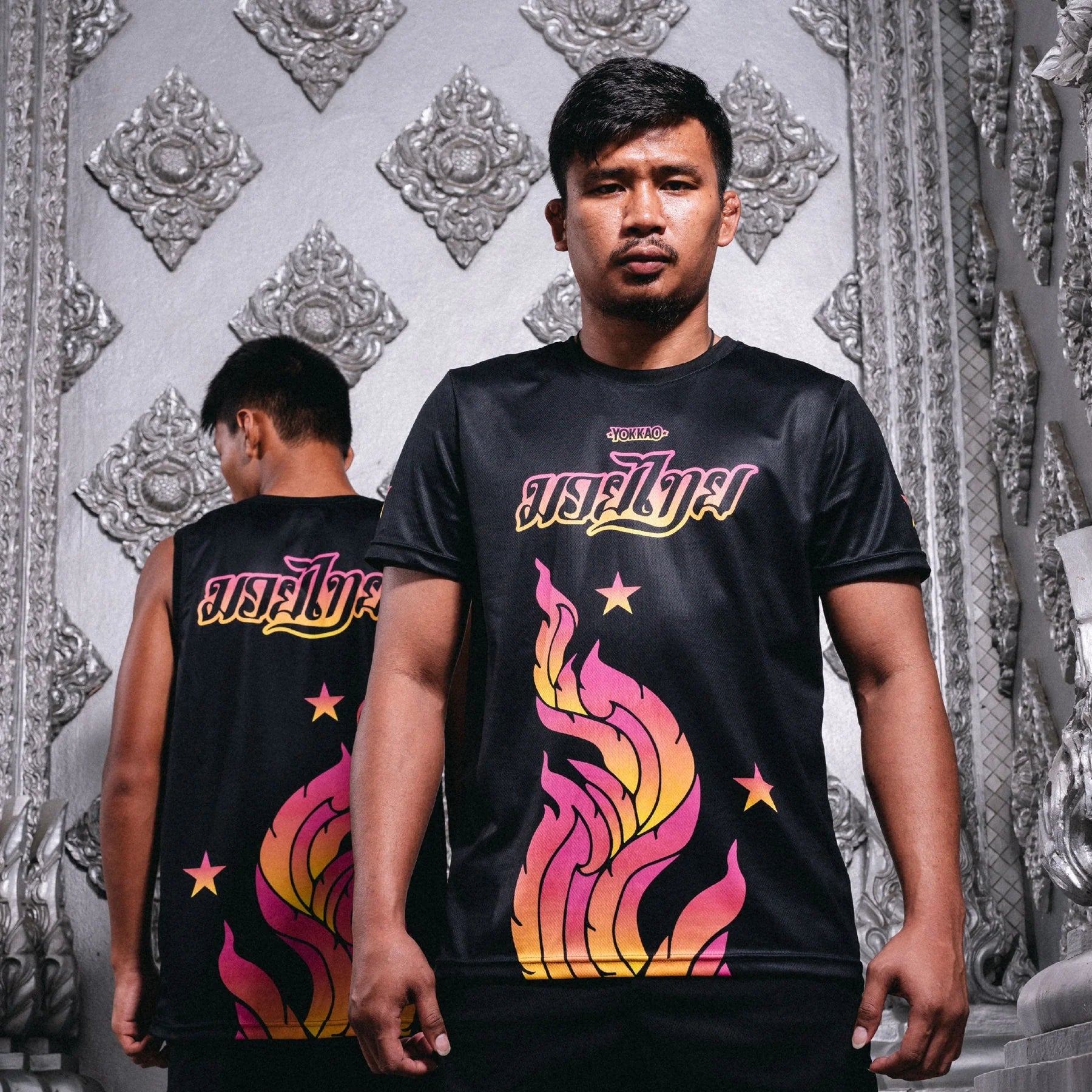 Man wearing a black t-shirt with colorful graphics and text, standing in front of a decorative silver wall.