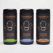 Three black cylindrical canisters of Champion Wipes biodegradable cleaning wipes arranged side by side on a light gray background. The left canister is labeled 'Lemon草ase with Tea Tree Oil' with a green band, the middle is 'Fresh Lemonade with Tea Tree Oil' with a blue band, and the right is 'Unscented with Tea Tree Oil' with a brown band.