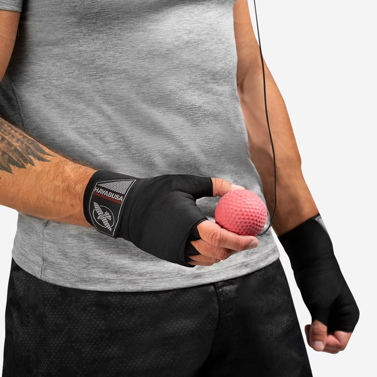 Person wearing black boxing hand wraps holding a pink exercise ball, with a gray shirt and black pants.