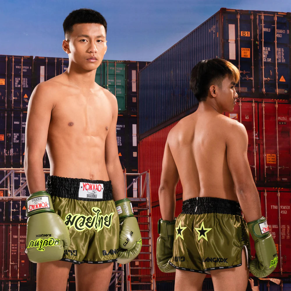 Two men wearing boxing shorts and gloves standing in front of shipping containers.