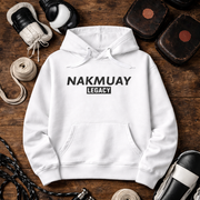 White hoodie with 'NAKMUAY LEGACY' text on a wooden surface with boxing equipment.