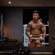 Boxer in red and yellow shorts standing in a boxing ring with gym equipment in the background.